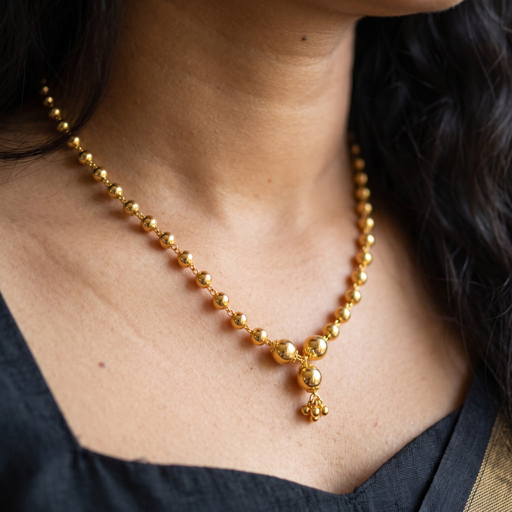 A close-up of a woman wearing a gold beaded necklace with a small hanging charm, shown against her neck and upper chest.